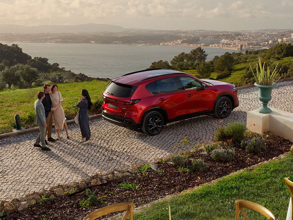 Rear three-quarter profile of a Soul Red Crystal Mazda CX-5 with a woman standing near the back, and a distant town visible across a body of water in the background.