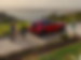 Profil arrière de trois quarts d'un Mazda CX-5 rouge vibrant cristal, avec une femme debout derrière le véhicule, et une ville lointaine visible au-delà d'une étendue d'eau en arrière-plan.