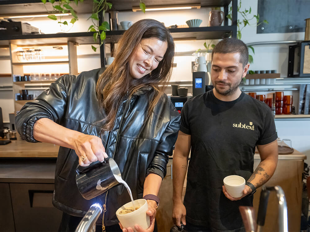A woman creates latte art in a coffee cup in front of a coffee shop employee.