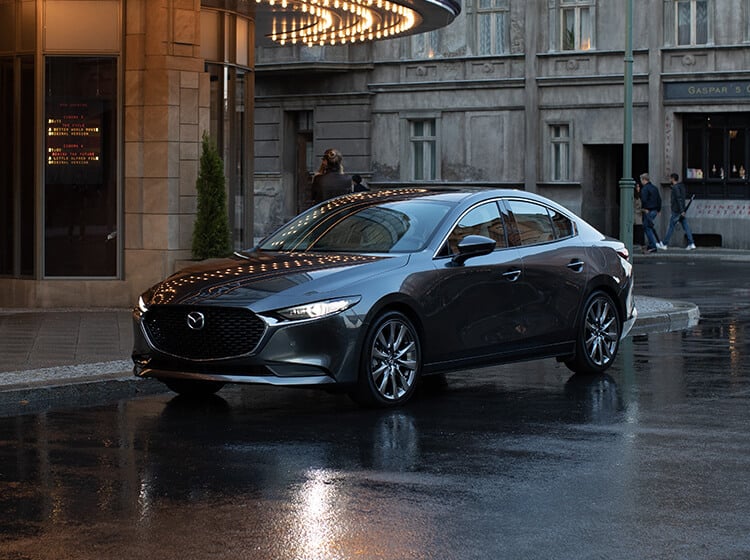 Mazda3 bleue les phares allumés, stationnée devant un hôtel, le soir, sur une chaussée mouillée
