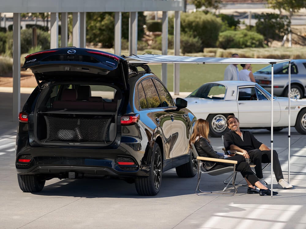 Two people sit on chairs next to a parked CX-90 with its trunk door open.