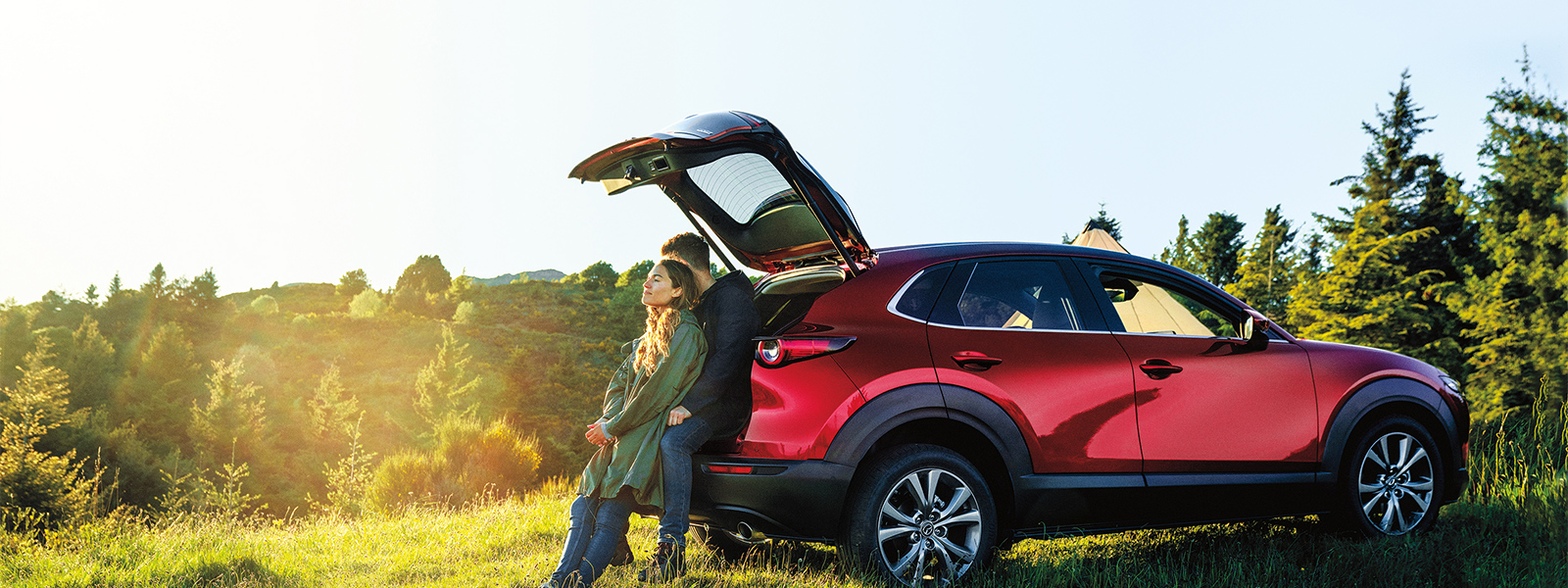 Jeune femme penchée vers un jeune homme assis à l’arrière d’un VUS Mazda rouge, le hayon ouvert, tous deux admirant la vue sur un paysage champêtre vallonné 
