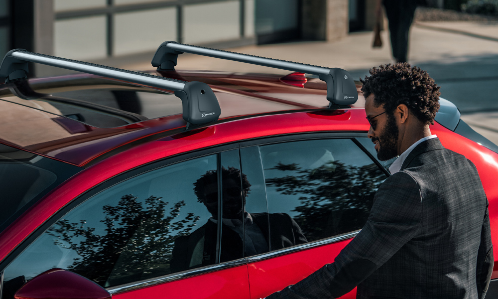 Homme en veston se penchant pour ouvrir la porte côté conducteur d’une Mazda3 Sport rouge, avec des accessoires sur le porte-bagages de toit