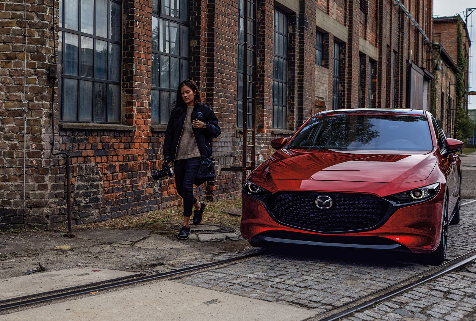 Woman walking alongside red Mazda3 parked on cobblestone next to red-brick warehouse.