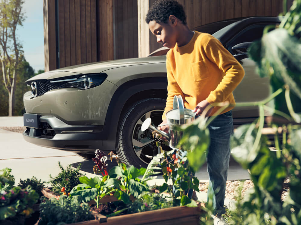 Jeune garçon arrosant son jardinet, avec un véhicule Mazda à l’arrière-plan