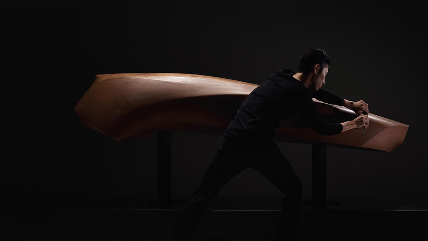 Un artisan de Mazda habillé en noir, tenant un outil dans chaque main, façonne une voiture en argile brune.