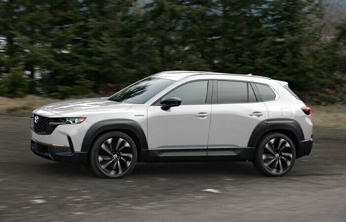 A white CX-50 Hybrid drives alongside trees