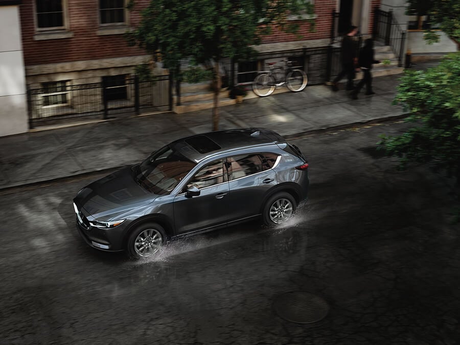 Vue vers le bas côté conducteur d’un véhicule Mazda gris sur une rue urbaine sous une forte pluie.