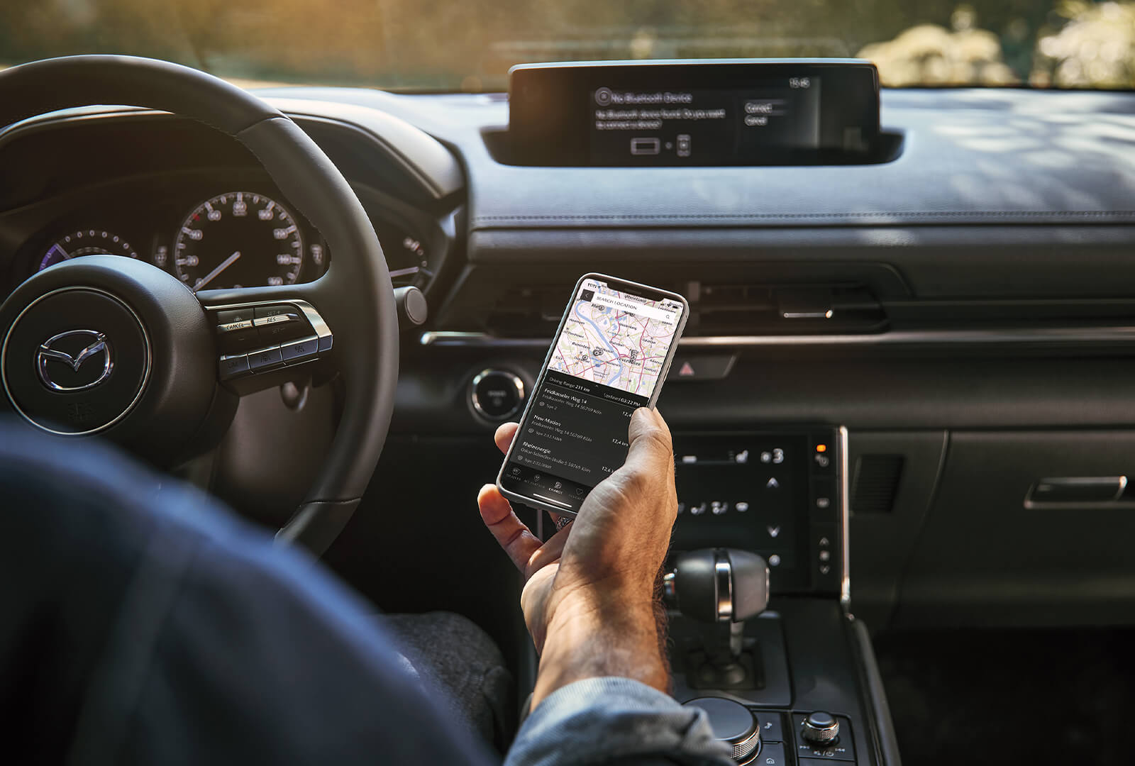 A man in the driver’s seat holds a smartphone displaying a map within the MyMazda app. 
