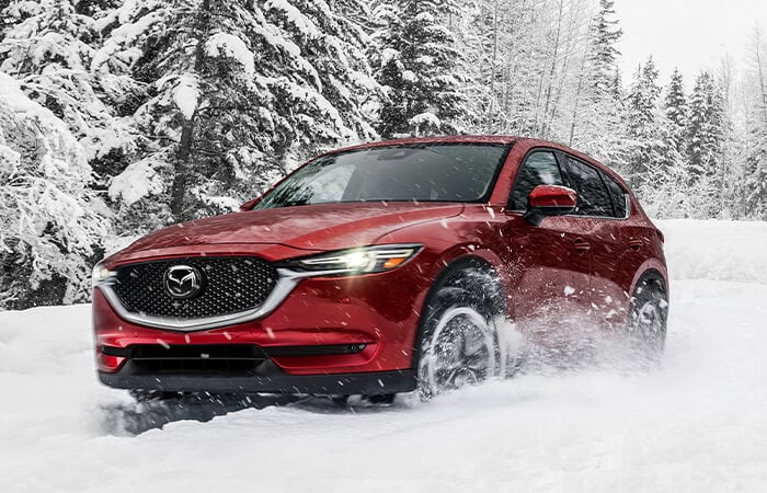 Soul Red Crystal Mazda SUV with headlights on drives past snow-covered conifers as it negotiates a curve in deep snow on a rural road. 
