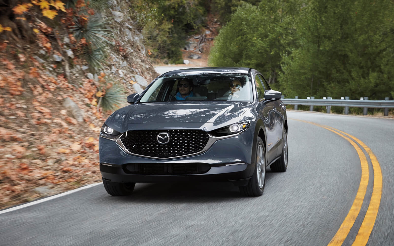Grey Mazda CX-30 SUV drives uphill curve in hill country 