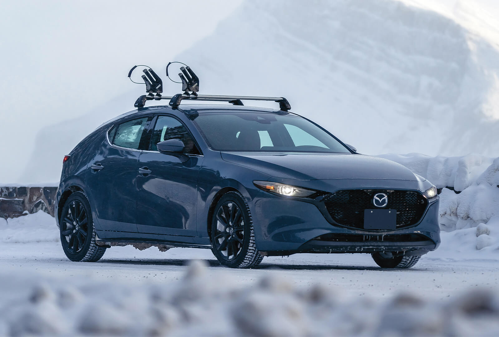 Blue Mazda3 Sport with ski rack parked against snow-covered mountain backdrop 