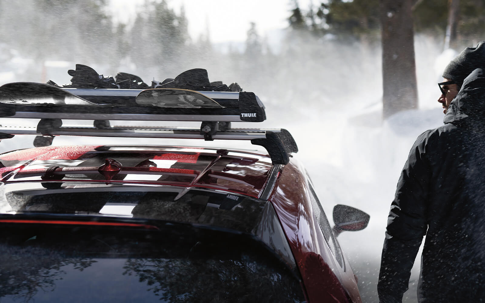 Personne en parka marchant à côté d’une Mazda rouge avec des snowboards sur le toit et un paysage enneigé au loin. 