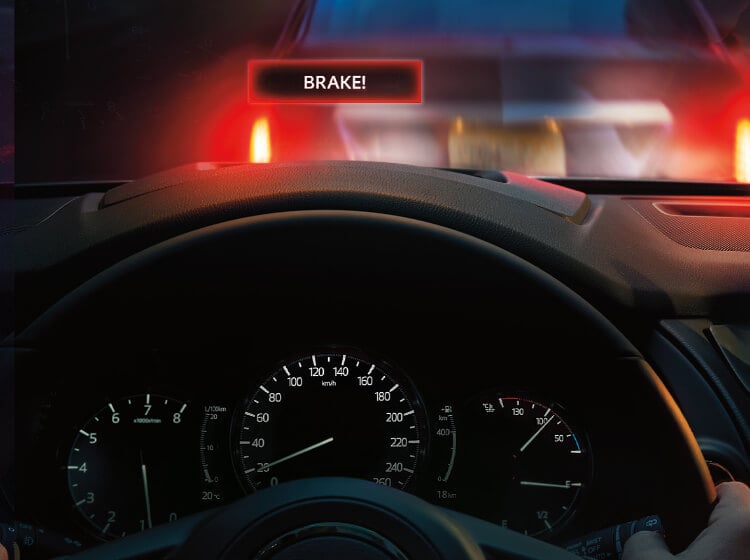 Vue d'un tableau de bord avec le mot « BRAKE! » en lettres blanches sur un rectangle noir avec un contour rouge apparaissant sur le pare-brise alors que l’arrière de la voiture qui précède occupe tout le champ de vision. 