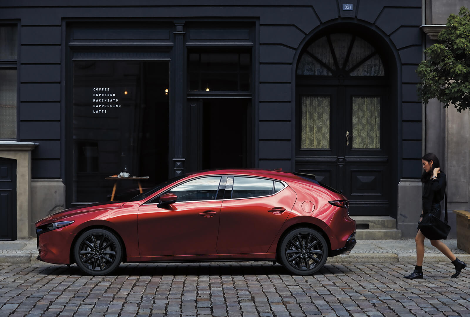 Une Mazda3 rouge vue de côté, dans une rue pavée, avec une jeune modeuse qui s’approche depuis la droite.