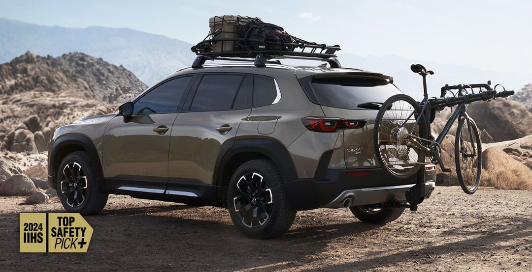 Vue arrière du Mazda CX-50 sable de zircon métallisé stationné, avec le porte-bagages de toit chargé d’équipements de plein air et un vélo de montagne sur le porte-vélos.