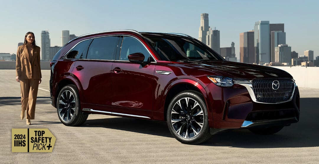 Vue de trois quarts avant d’un Mazda CX-90 hybride léger rouge artisan stationné, avec un panorama urbain à l’arrière-plan. Une femme marche à côté du véhicule.