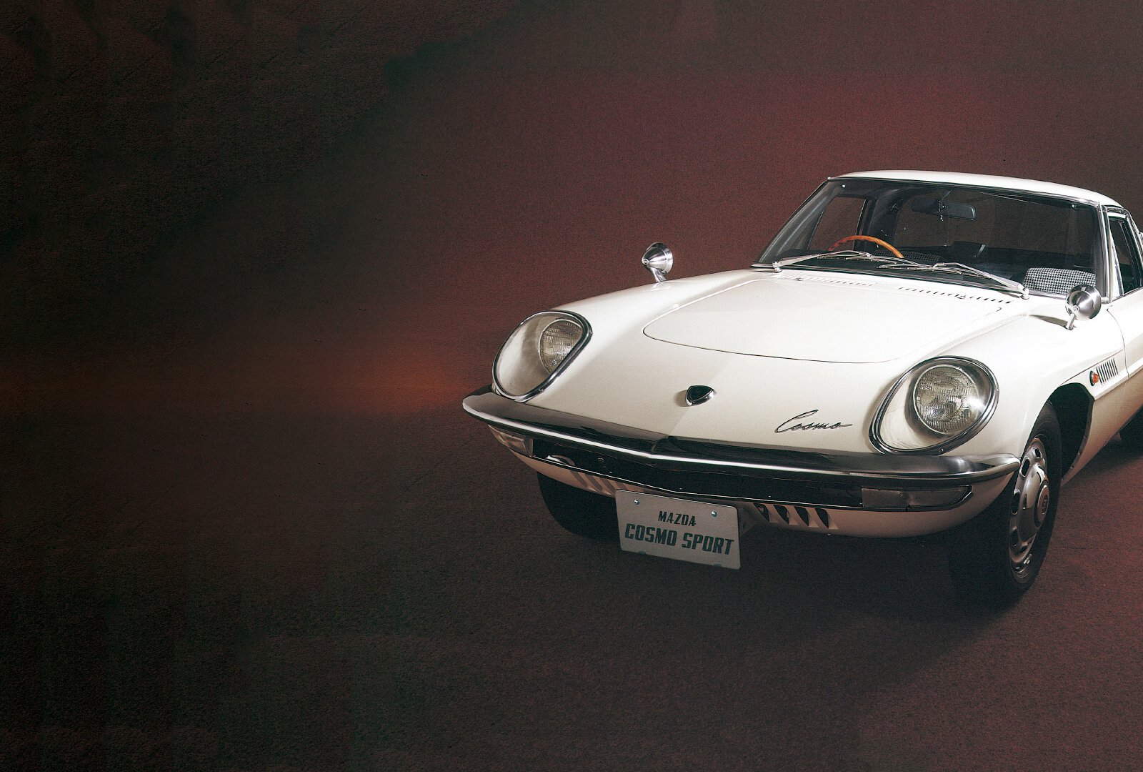 White Mazda Cosmo Sport against red background in three-quarter view from in front of passenger-side headlight.