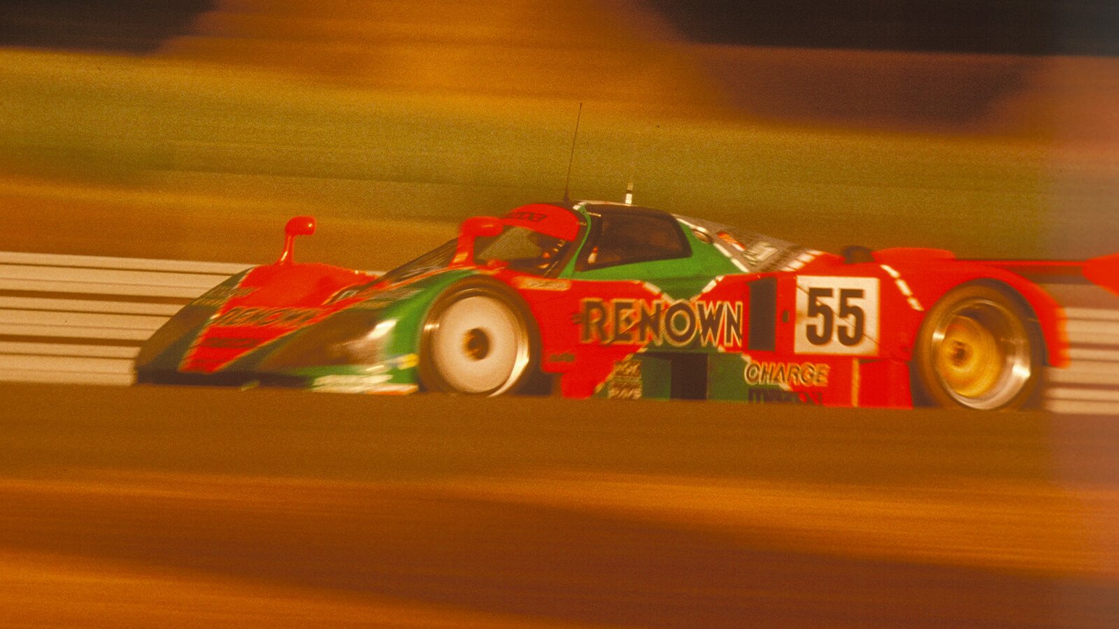 Mazda 787B No.55 racing car in red and green trim against a blurred background races at the Le Mans 24-Hour Endurance Race.