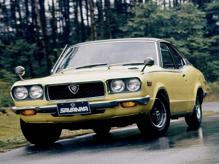 Yellow Mazda RX-2 on wet road with blurred trees in background viewed from in front of driver-side headlight