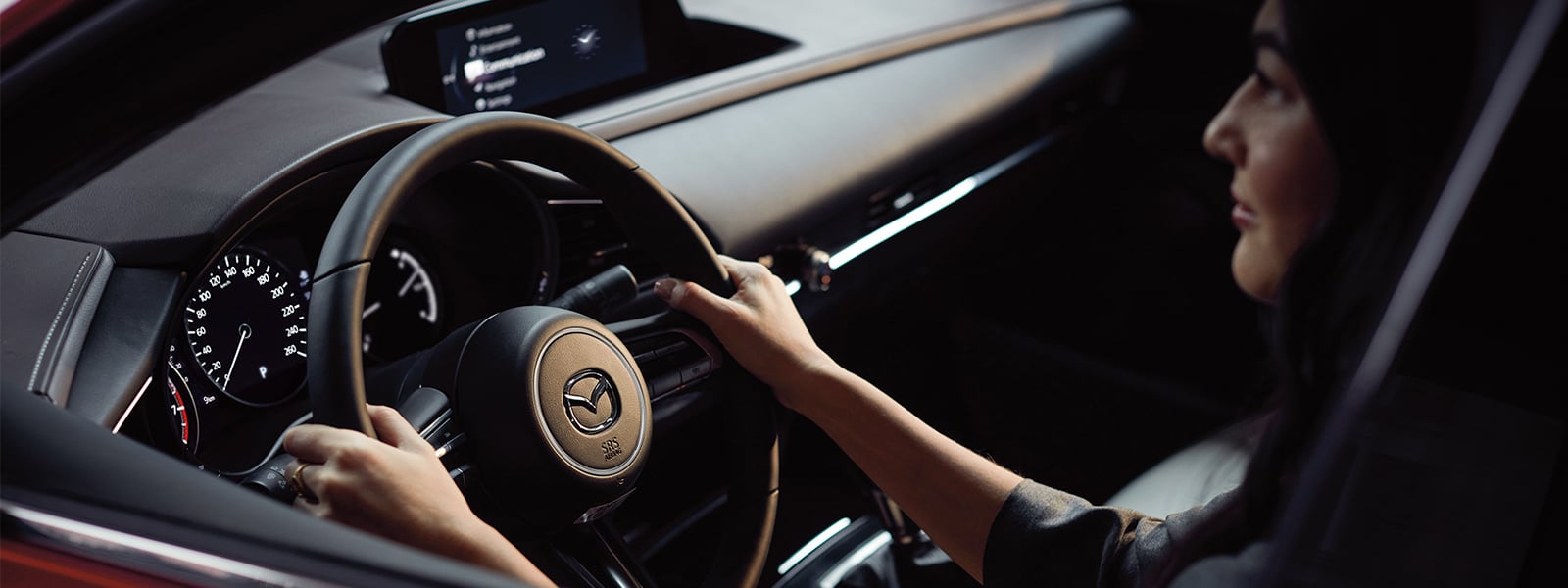 Interior over-the-shoulder shot of woman with hands at 10 and 2 on the wheel with the Mazda logo catching the light.
