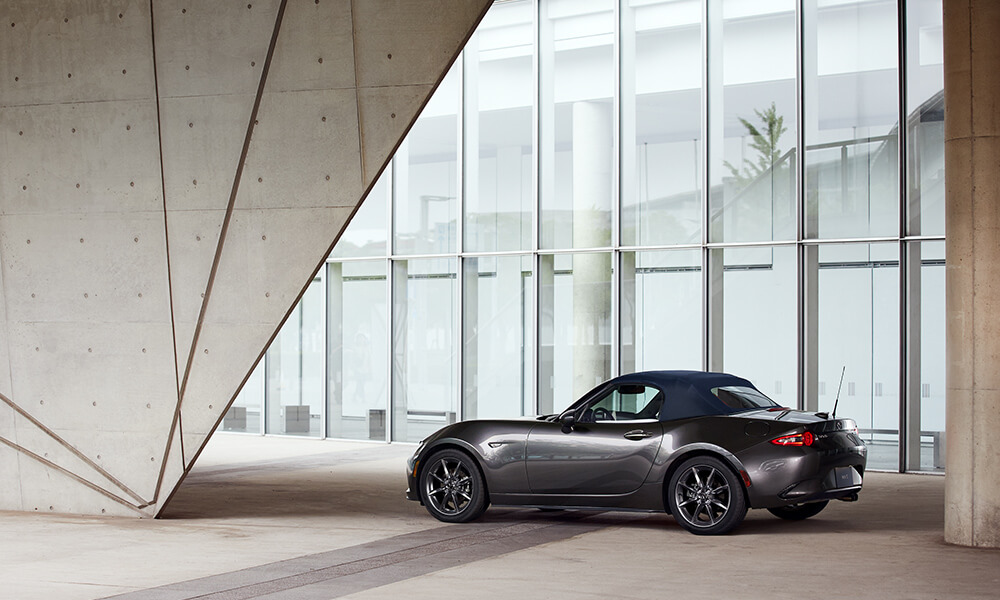 Machine gray metallic Mazda MX-5 Soft Top 2-Seat Convertible between concrete pillars and in front of retailer showroom window.