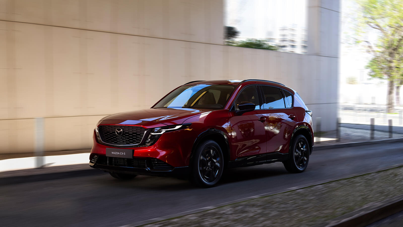 Rear side profile of a Soul Red Crystal Mazda CX-5, partially visible up to the back wheel, parked in a shadowed interior driveway, framed by an open view of distant mountains.