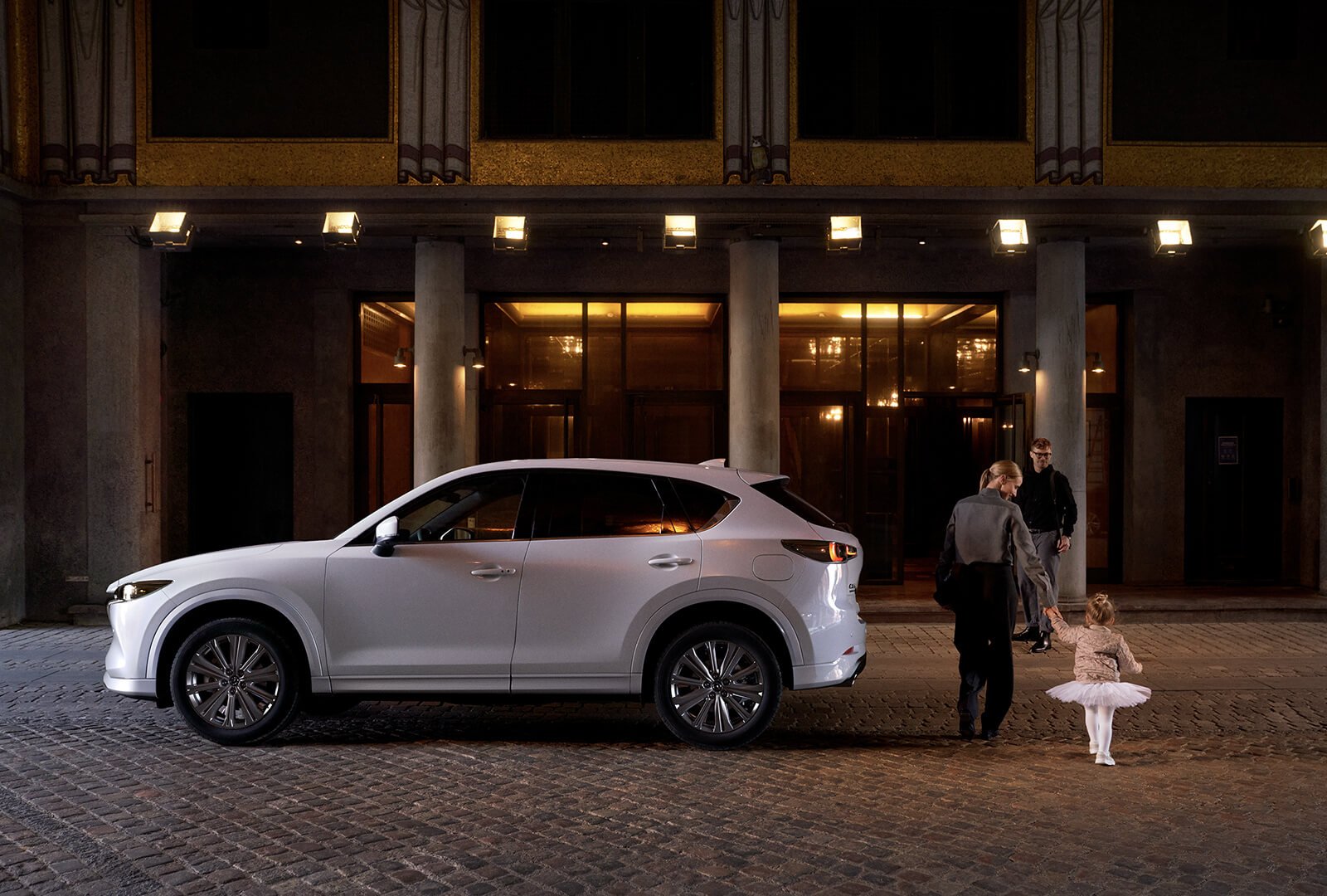 Un Mazda CX-5 blanc stationné, vu de profil, alors qu’une maman accompagne sa fillette en tutu blanc, en passant derrière le véhicule et en se dirigeant vers le papa qui attend devant l’entrée d’un hôtel votre Mazda.