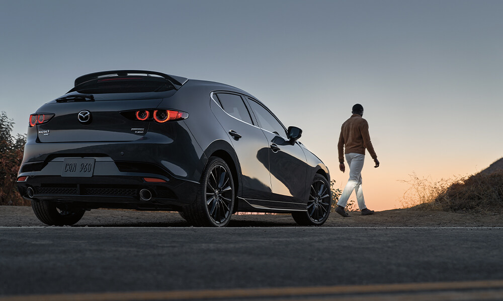 Jet Black Mica Mazda3 Sport parked on side of highway as  owner walks towards trail with a sunset view.