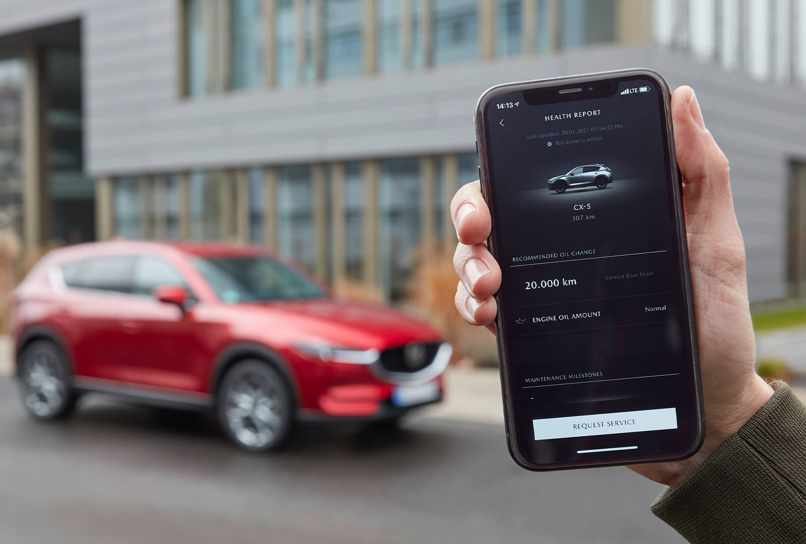 Closeup, hand holds smartphone displaying CX-5 vehicle health report with Crystal Red Metallic CX-5 in the background.  