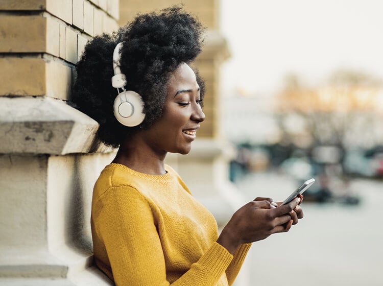 Smiling woman with headphones looking at her smartphone while leaning against a brick wall