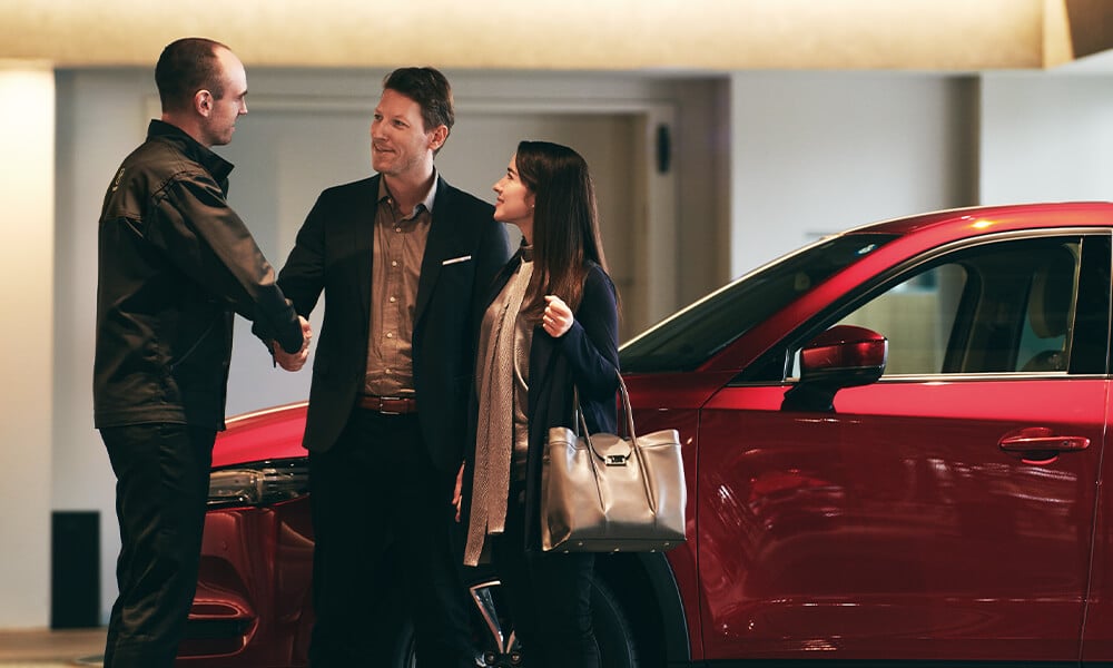 Couple bien habillé souriant qui serre la main d’un représentant Mazda, alors qu’ils sont devant un véhicule Mazda rouge.