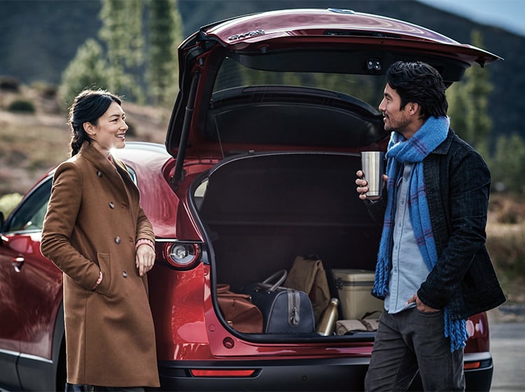 Woman in brown coat and man with scarf and coffee talking in front of open hatch of red Mazda3 parked in the outdoors. 