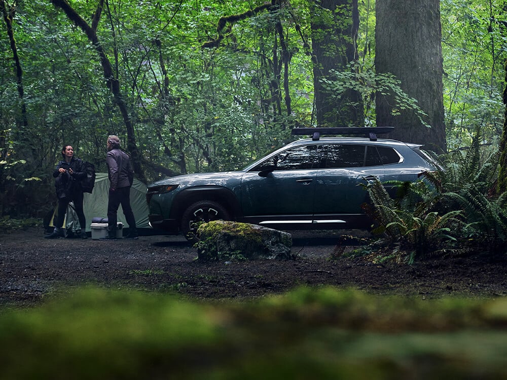Un couple discute entre une tente et un Mazda CX-50 garé au milieu d'une végétation luxuriante et de troncs d'arbres.