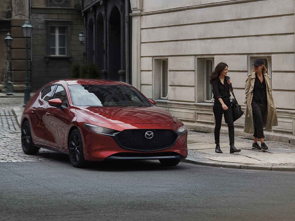 Soul Red Crystal Metallic Mazda3 Sport drives past two young women walking on sidewalk in historic district.