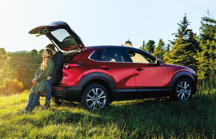 Couple sit in open hatch of Soul Red Crystal Metallic Mazda CX-30 parked in grassy field.