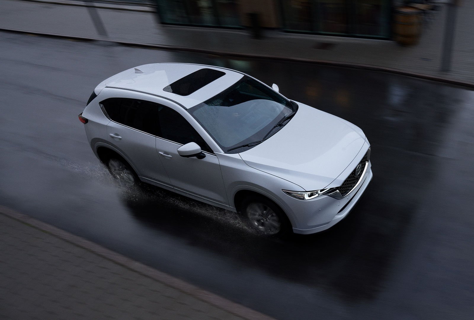 Overhead view of Snowflake White Pearl Mazda CX-5 with moonroof driving along city street in rain.