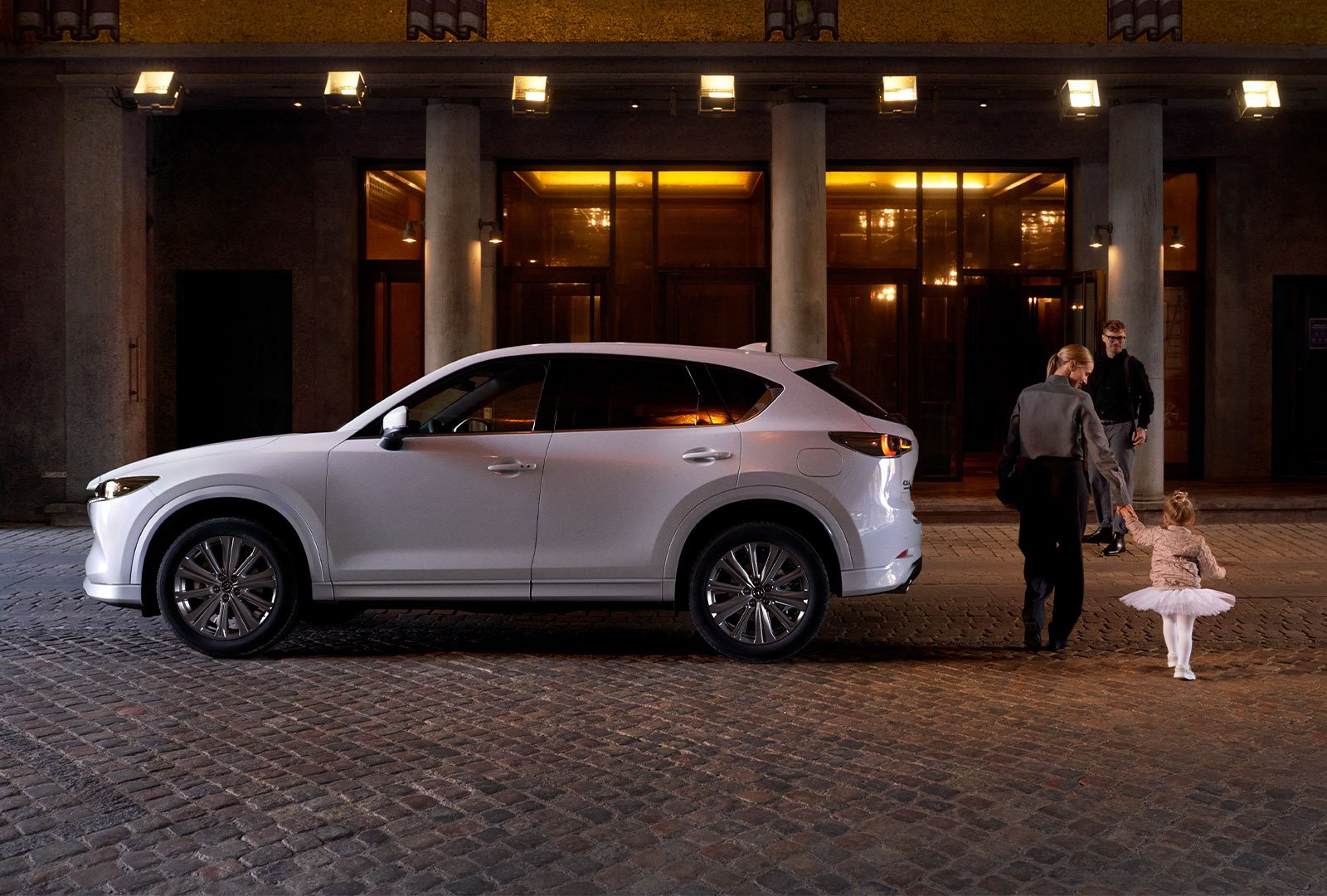 Une femme escorte une enfant en tutu blanc jusqu’à l’entrée d’un bâtiment, devant un Mazda CX-50 blanc rhodium métallisé. 