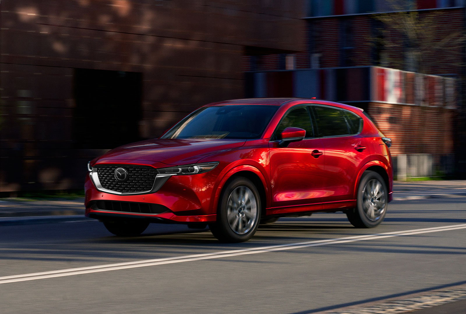 Un Mazda CX-5 rouge vibrant cristal métallisé passe devant des structures floues dans une rue.