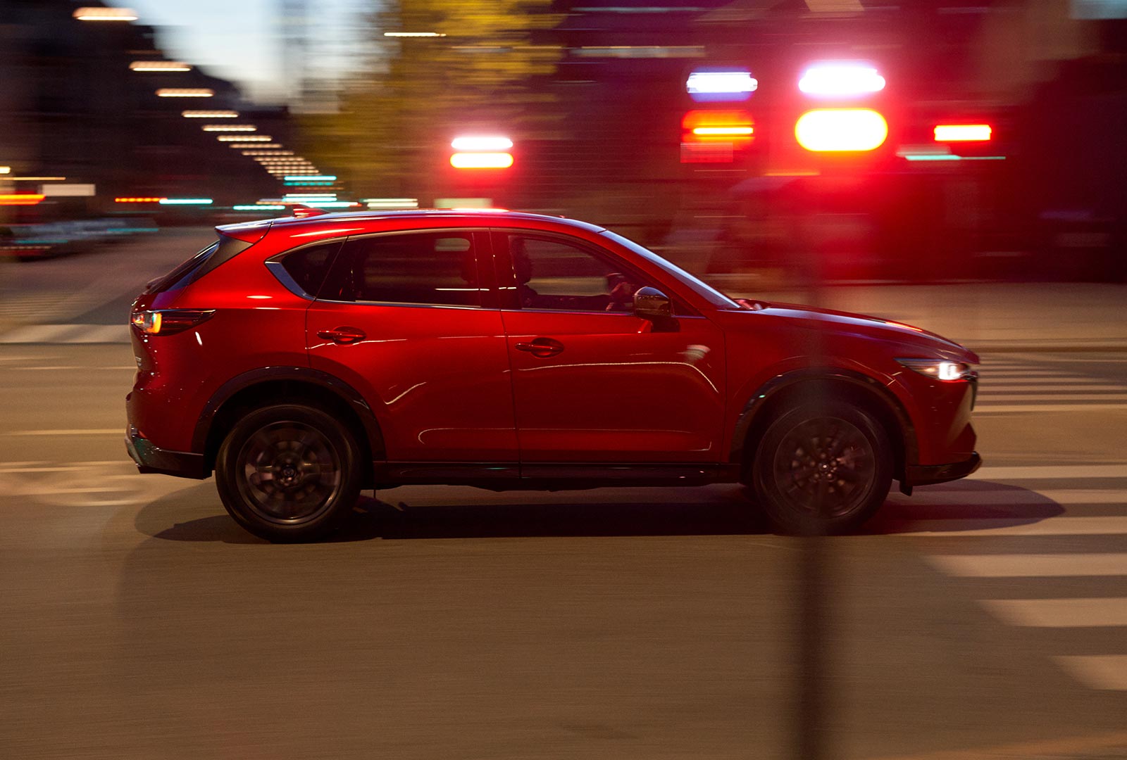 Soul Red Crystal Metallic Mazda CX-5 crosses intersection with blurred traffic lights at twilight.