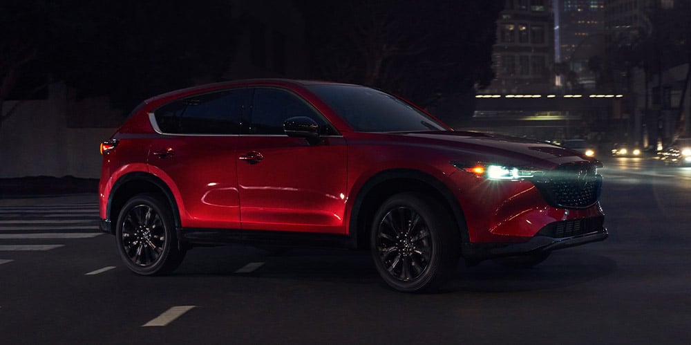 Un Mazda CX-5 Design sportif rouge vibrant cristal métallisé avec les phares allumés traverse une intersection de nuit.  
