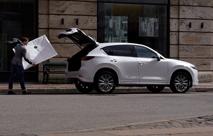 Man carries white ? to Snowflake White Pearl Mazda CX-5 parked on city street with liftgate open.