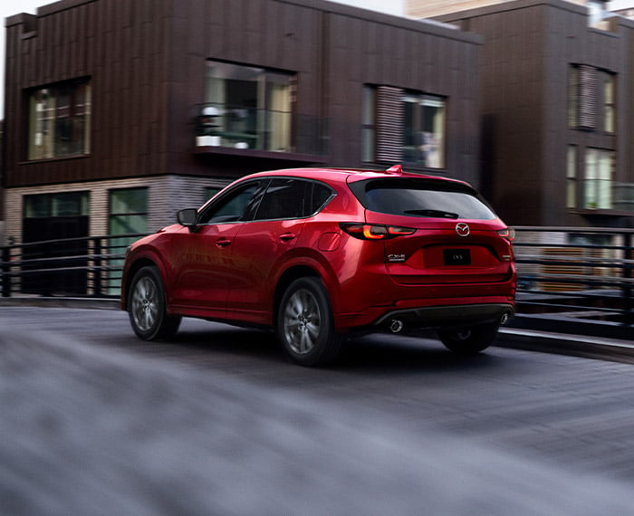 Un Mazda CX-5 rouge vibrant cristal métallisé s’éloigne sur un viaduc, devant des bâtiments de couleur marron.