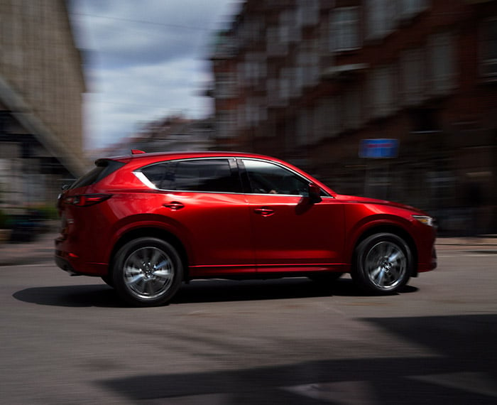 Une Mazda CX-5 rouge vibrant cristal métallisé traverse une intersection ensoleillée.