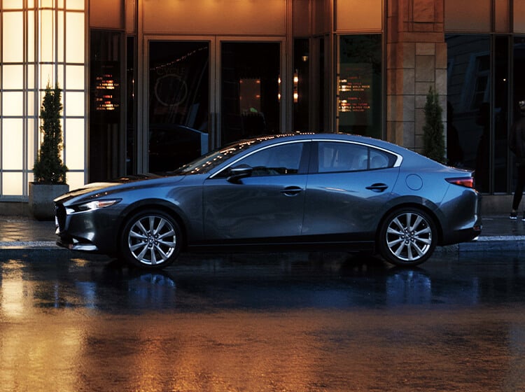 Une berline Mazda3 de couleur quartz platine métallisé garée devant l’entrée d’un hôtel au crépuscule.