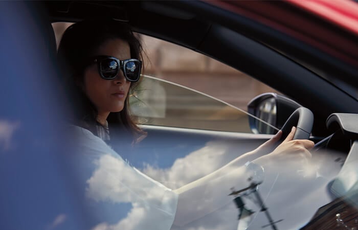 Female driver wearing sunglasses seen through passenger side window.