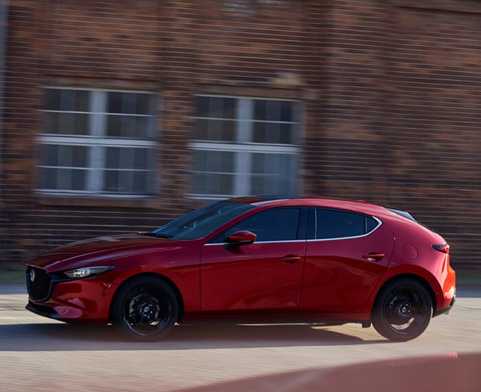 Soul Red Crystal Metallic Mazda3 Sport hatchback drives past blurred brick building.