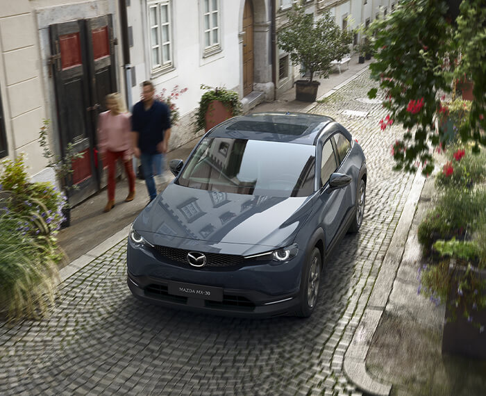 Overhead view of Polymetal Grey MX-30 EV driving up narrow cobblestone street flanked by planters.