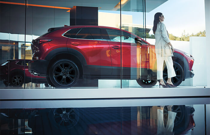 Driver standing next to Soul Red Crystal Metallic CX-30 in parking spot encased in glass.