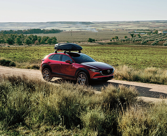 Soul Red Crystal Metallic Mazda CX-30 with roof storage travels down farm land dirt road.